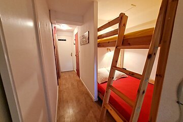 A narrow hallway with a wooden floor leads to doors, adjacent to a cozy bunk bed alcove featuring red and yellow bedding.