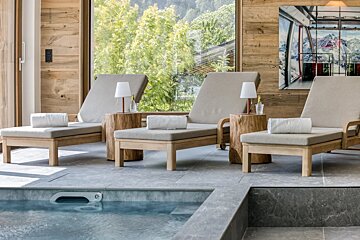 A cozy indoor spa with three wooden lounge chairs next to a pool, featuring large windows with forest views and rustic decor.