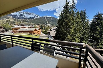 Balcony view of a mountain resort with wooden chalets, pine trees, and snowy peaks.