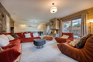 Cozy ski lodge living room with plush red sofas, a warm fireplace, rustic wood/stone walls, and large windows revealing snowy views.