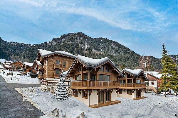 Snow-covered wooden chalets in a winter mountain village, with pine forests and a blue sky in the background.