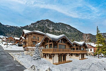 Snow-covered wooden chalets in a winter mountain village, with pine forests and a blue sky in the background.