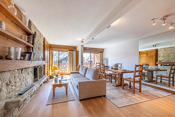 A warm, rustic open-plan living and dining room with a stone fireplace, wooden furniture, and mountain views from two balconies.