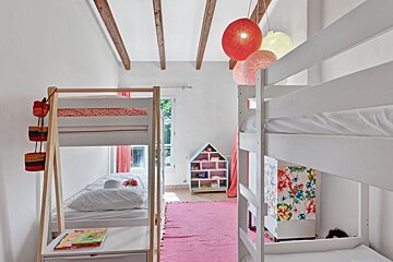 A colorful, bright kids' room with two bunk beds, a pink rug, hanging lanterns, exposed beams, and a dollhouse bookshelf.