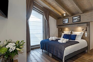Cozy alpine-style bedroom with exposed wooden beams, a bed with blue accents, and a large window.