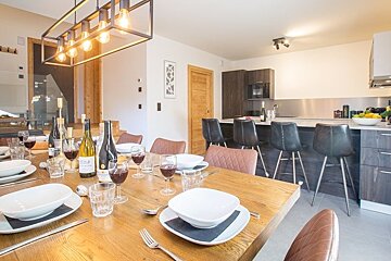 A set wooden dining table with wine and bowls in a modern open-plan kitchen and dining area, featuring a stylish hanging light fixture.