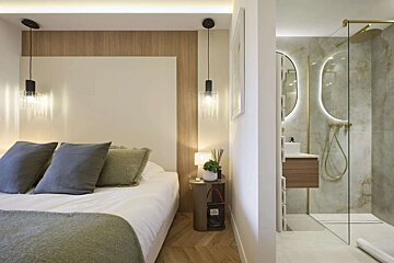 A modern bedroom with a bed, wooden paneling, and pendant lights, opening to a chic bathroom with a glass shower and backlit vanity mirror.