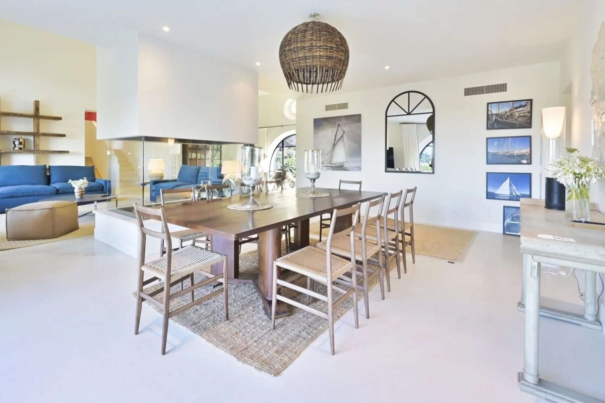 Spacious, modern dining and living room with a long table, blue sofas, glass fireplace, and nautical art. Features a woven pendant light.