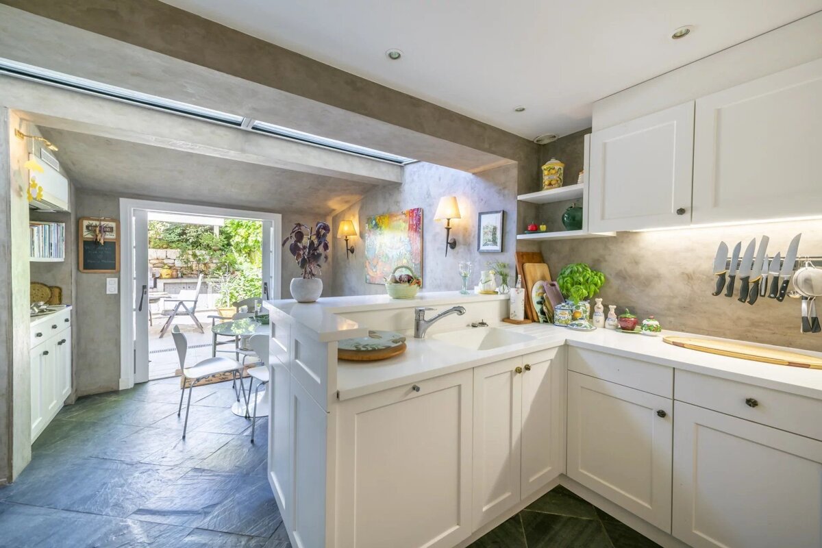 A bright, modern kitchen with white cabinets, grey concrete walls, and slate floors, opening to an outdoor patio.