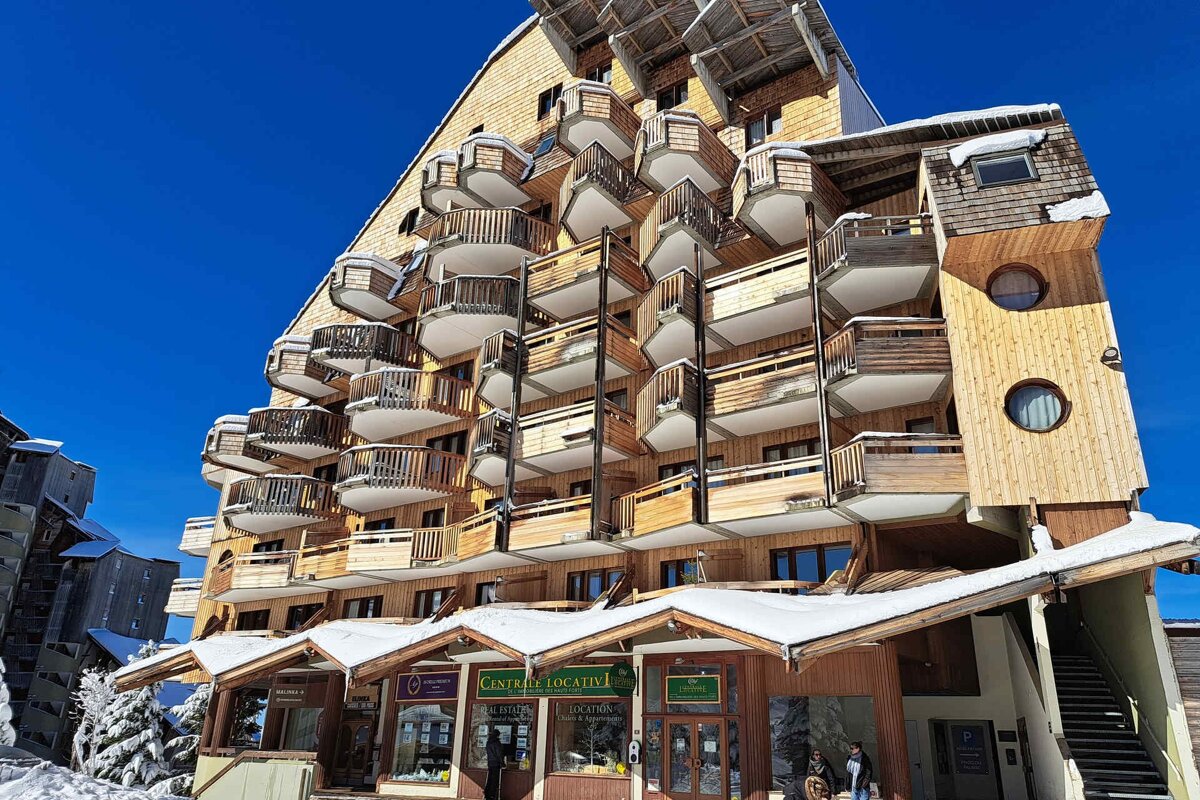 A unique, multi-story wooden chalet building with numerous balconies, dusted with snow, under a bright blue sky. Shops line the ground floor.