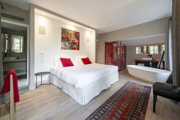 Stylish open-concept bedroom featuring a large bed with red pillows, an in-room freestanding bathtub, and light wooden floors.