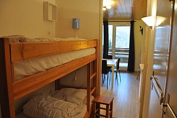 Wooden bunk beds in a cozy room with a view of a dining table and balcony in the background.
