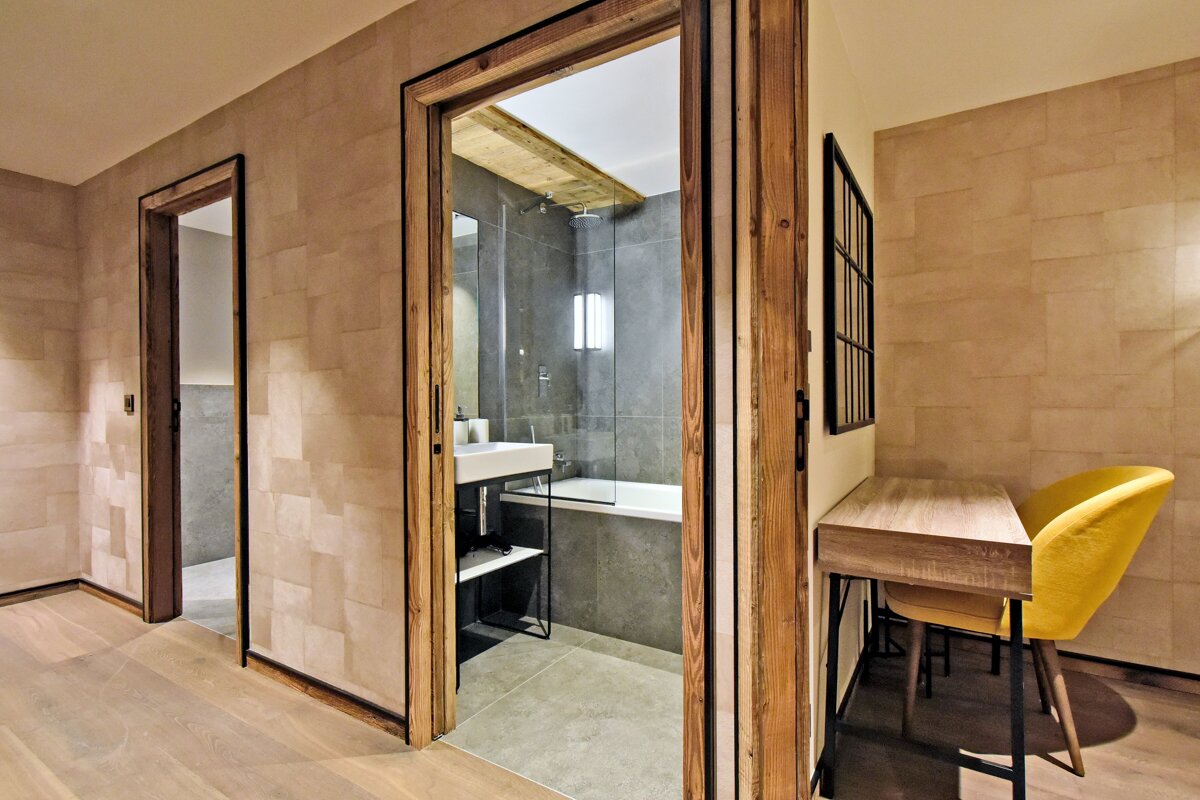 Modern hallway with two open wooden doorways. One reveals a stylish bathroom with a tub, shower, and sink. A desk and yellow chair are to the right.