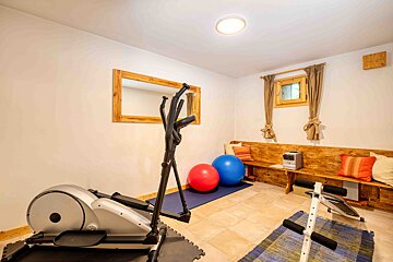 A bright home gym with an elliptical, exercise balls, ab bench, and a wooden bench with cushions. A mirror and window are on the wall.
