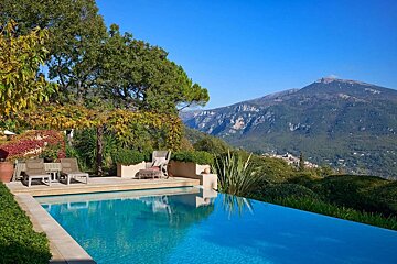 A luxurious infinity pool overlooks a breathtaking mountain landscape and lush garden under a clear blue sky, with lounge chairs for relaxation.