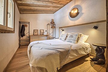 Cozy, rustic bedroom with a white bed, wooden ceiling and floor, open wardrobe, and a unique bear-shaped side table. Warm lighting adds ambiance.