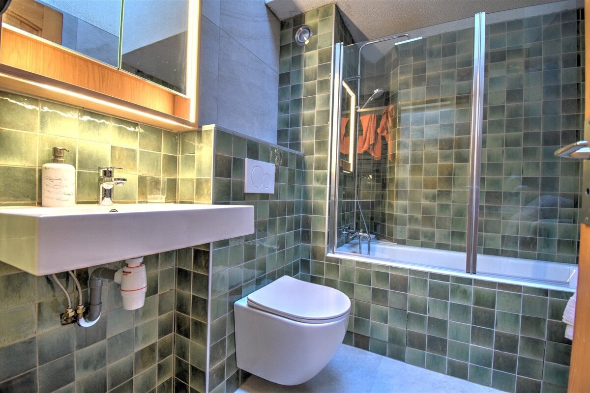 A modern bathroom with green square tiles, featuring a sink, wall-mounted toilet, and a bathtub with a glass shower screen.