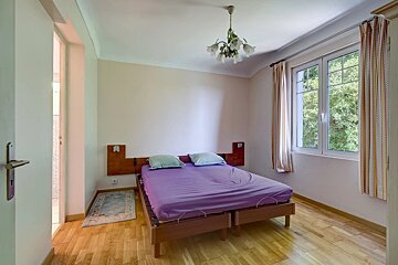 A simple bedroom featuring a purple bed, wooden floors, and a window looking out to green trees.