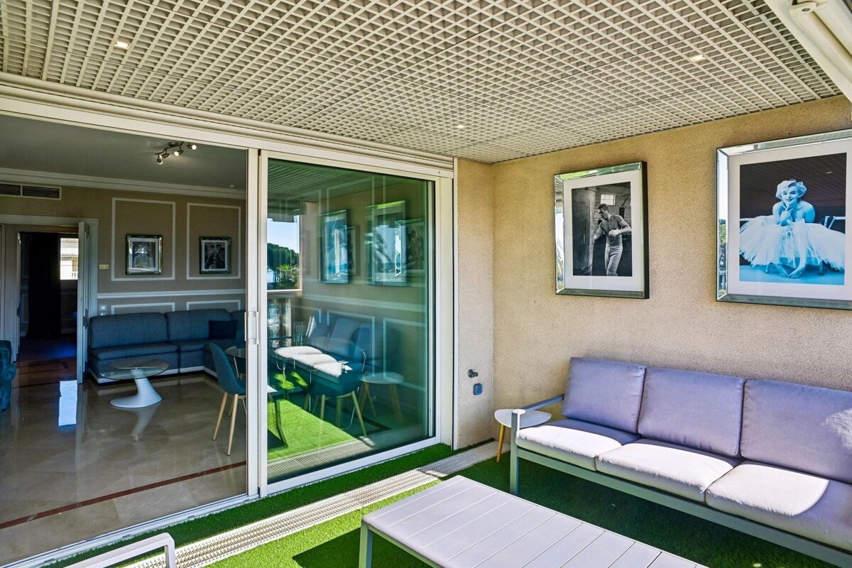 An outdoor terrace with artificial grass, a grey sofa, framed photos, and large sliding glass doors leading to a living room.