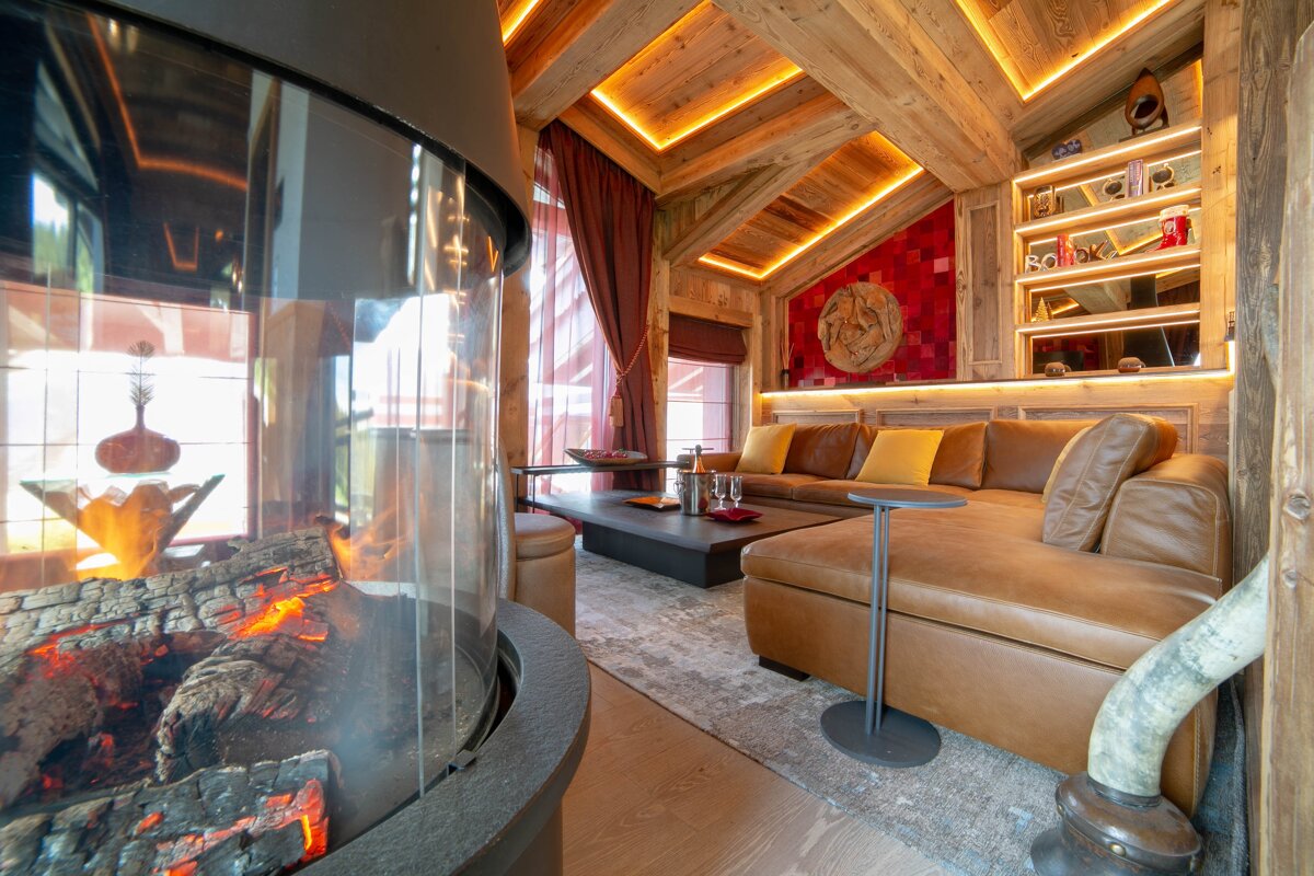 Cozy chalet lounge with a curved glass fireplace, a brown leather sectional, and warm wood-paneled walls with ambient lighting.