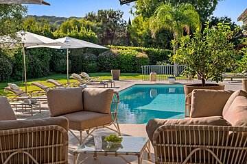 Elegant poolside lounge area with comfortable patio furniture, sun loungers, and umbrellas, set in a lush green garden under a clear sky.
