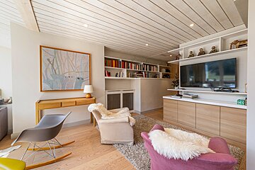 A modern living room with a slatted wood ceiling, a painting, built-in shelves, TV, and diverse seating including a rocker and armchairs with fur.