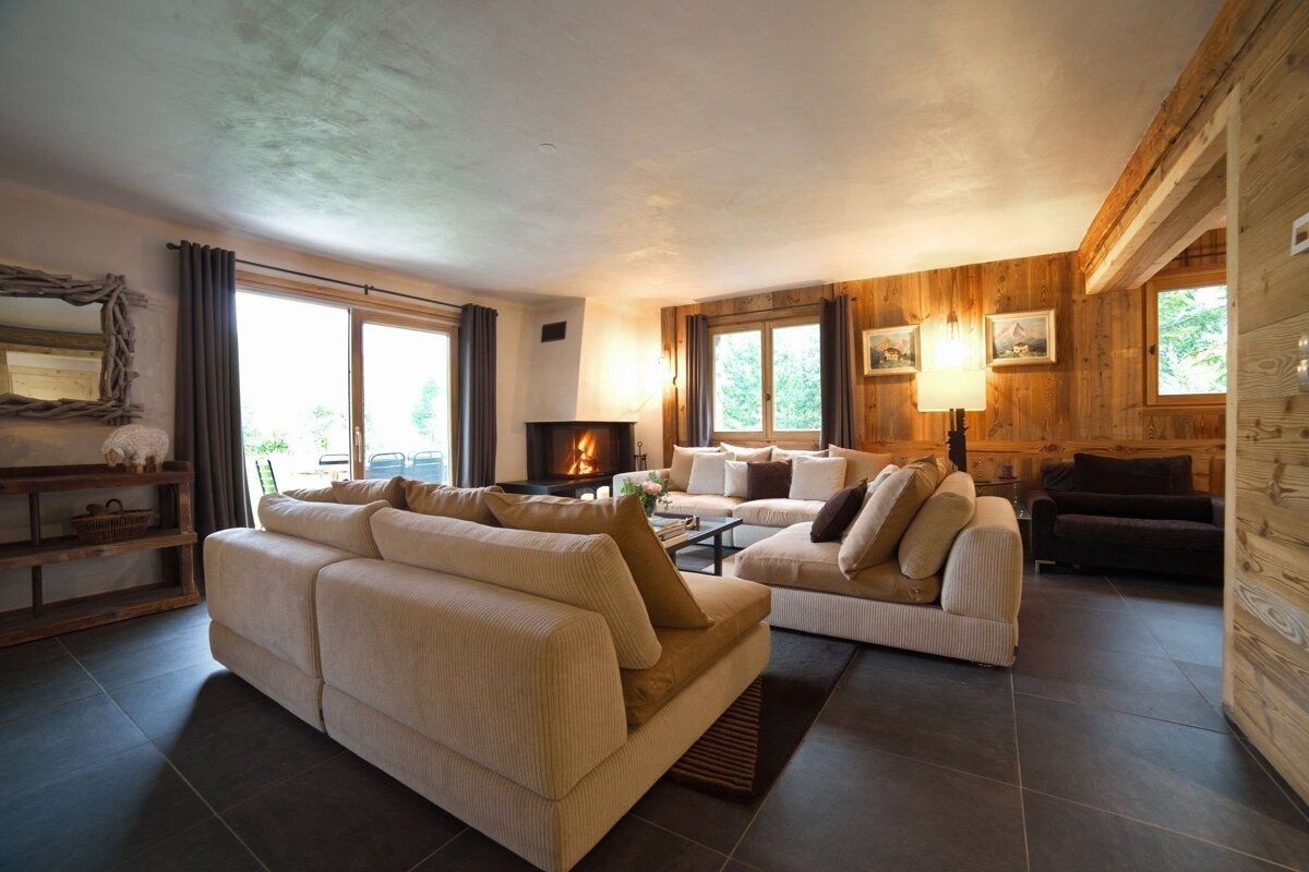 Cozy living room with comfy sectional sofas, a crackling fireplace, rustic wood walls, big windows, and dark tiled floors.