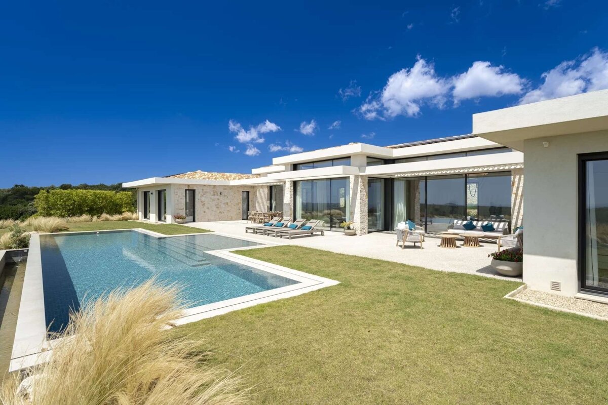 Modern white villa with a large swimming pool, green lawn, and outdoor lounge areas under a clear blue sky with sparse clouds.