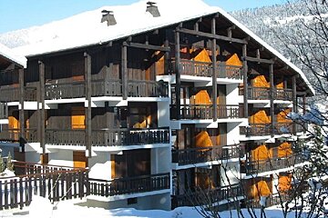 A large, multi-story wooden chalet apartment building with many balconies, covered in snow on a bright winter day.