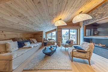 A luxurious wooden chalet living room featuring a long sofa, furry chairs, a sleek fireplace, and large windows offering a snowy mountain view.