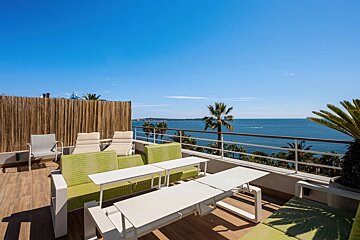 A sunny rooftop terrace with green couches, white tables, and lounge chairs overlooking a blue ocean.