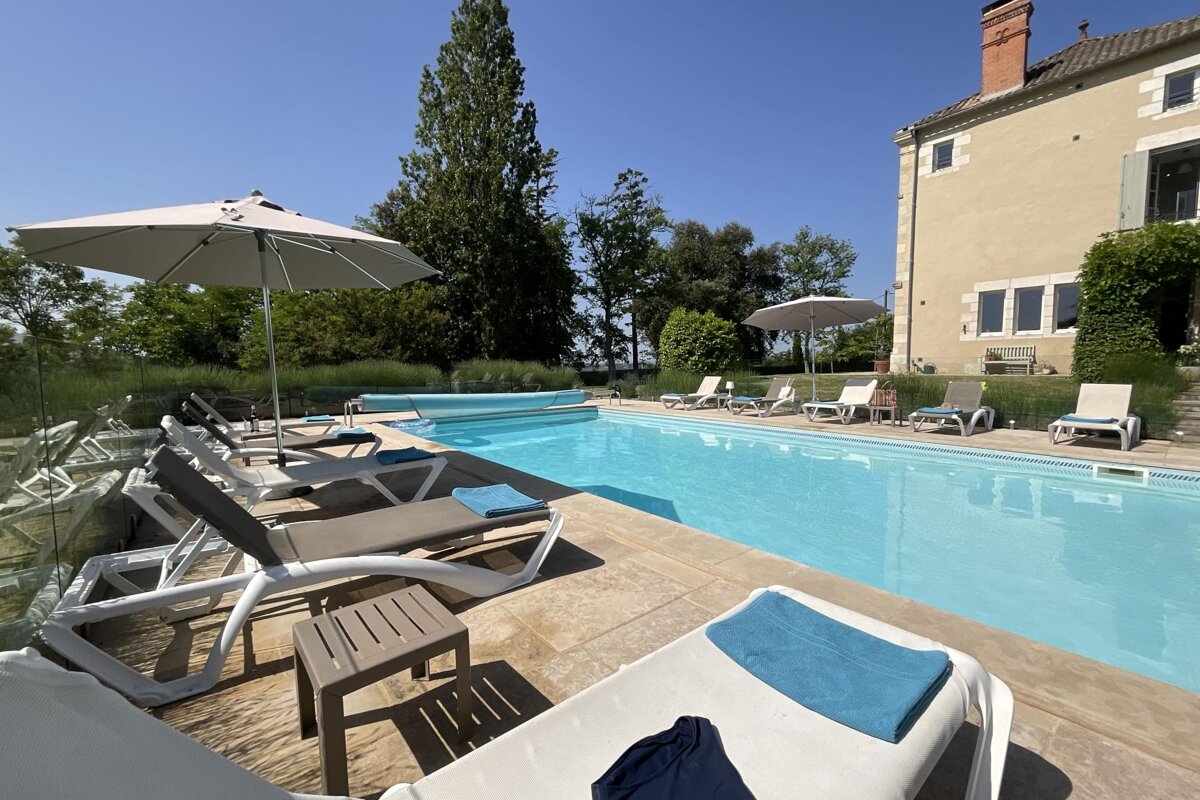 A beautiful outdoor swimming pool with sun loungers, umbrellas, and a large historic house under a clear blue sky.