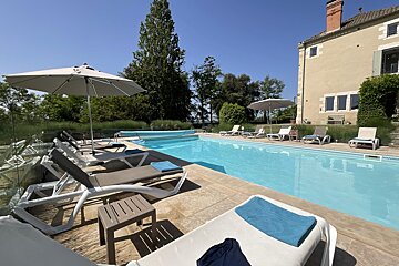 A beautiful outdoor swimming pool with sun loungers, umbrellas, and a large historic house under a clear blue sky.