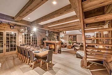 Luxurious wooden chalet with a large dining room, stone fireplace, cozy living area, and rustic staircase, all bathed in warm light.