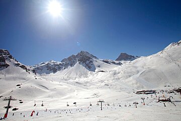 A sunny ski resort with expansive snow-covered mountains under a bright blue sky, featuring numerous skiers, chairlifts, and buildings.