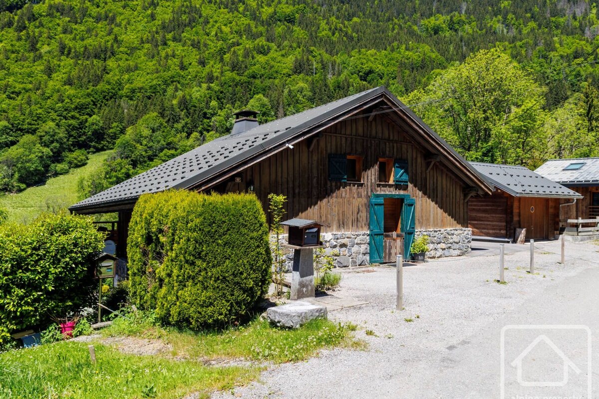 A rustic wooden chalet with blue shutters and a stone foundation, nestled against a lush green forested hillside. A gravel driveway leads to the front.