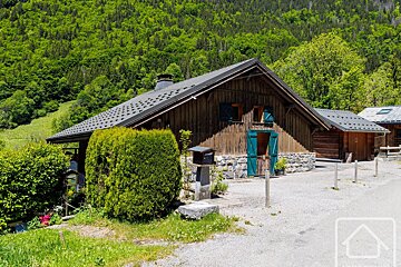A rustic wooden chalet with blue shutters and a stone foundation, nestled against a lush green forested hillside. A gravel driveway leads to the front.