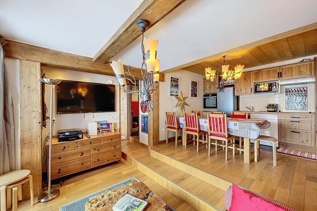 A cozy, split-level chalet interior featuring rustic wood accents, exposed beams, a living area with a TV, and an elevated dining/kitchen space.