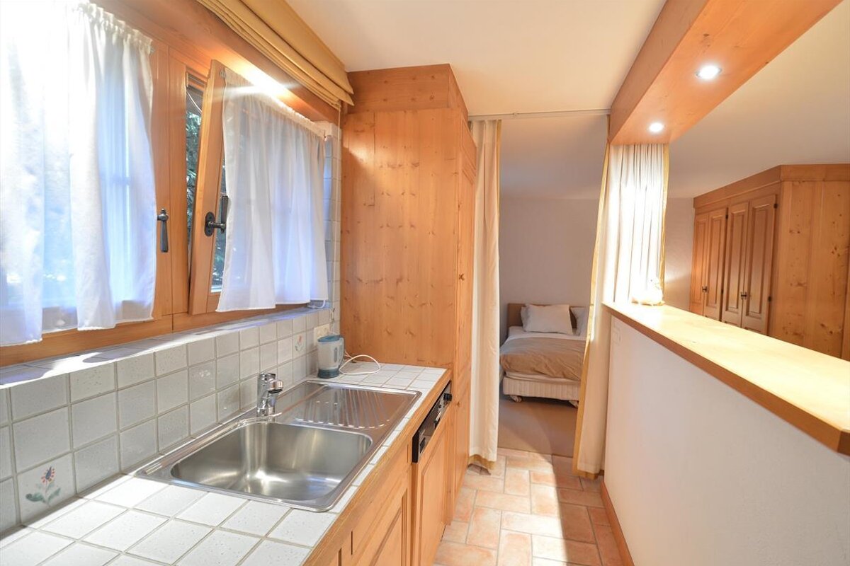 A bright studio apartment with a compact kitchenette (sink, tiled counter, window) adjacent to a curtain-separated bedroom, featuring warm wood finishes.