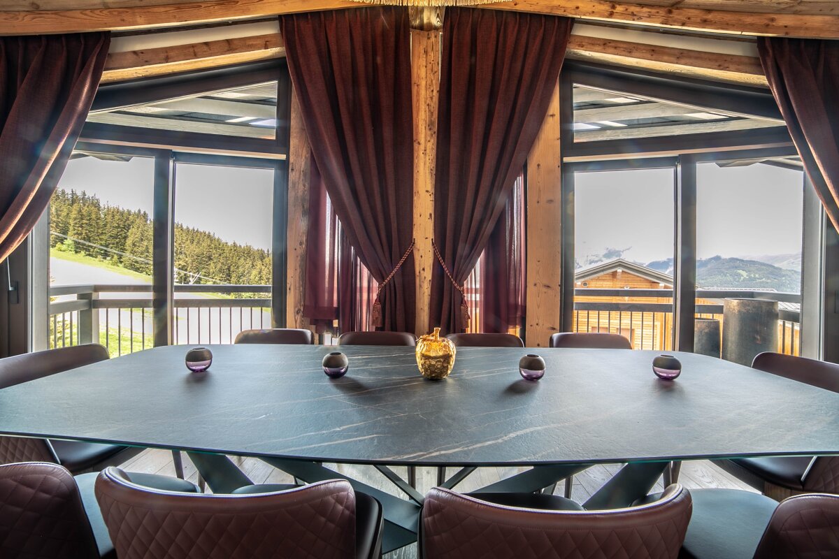 A stylish dining room with a large table, brown leather chairs, and expansive windows offering a panoramic mountain view with forests and a distant chalet.