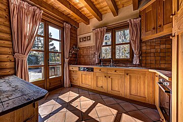 A cozy, rustic wooden kitchen featuring checkered curtains, tiled floors with sun patches, and natural light from large windows and a glass door.