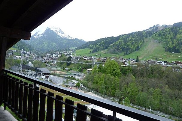 Balcony view of a lush green alpine valley with a village, forested hills, and a snow-capped mountain in the background.