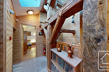 Rustic chalet interior with natural wood walls, exposed timber beams, and skylights. A hallway leads to a room, with a bookshelf and copper jugs.
