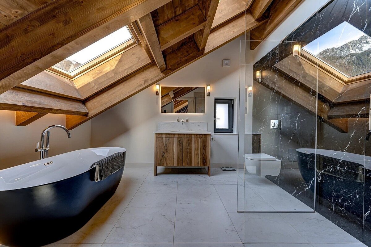 A luxurious attic bathroom features exposed wooden beams, a black freestanding tub, a marble shower, and skylights revealing snowy mountains.
