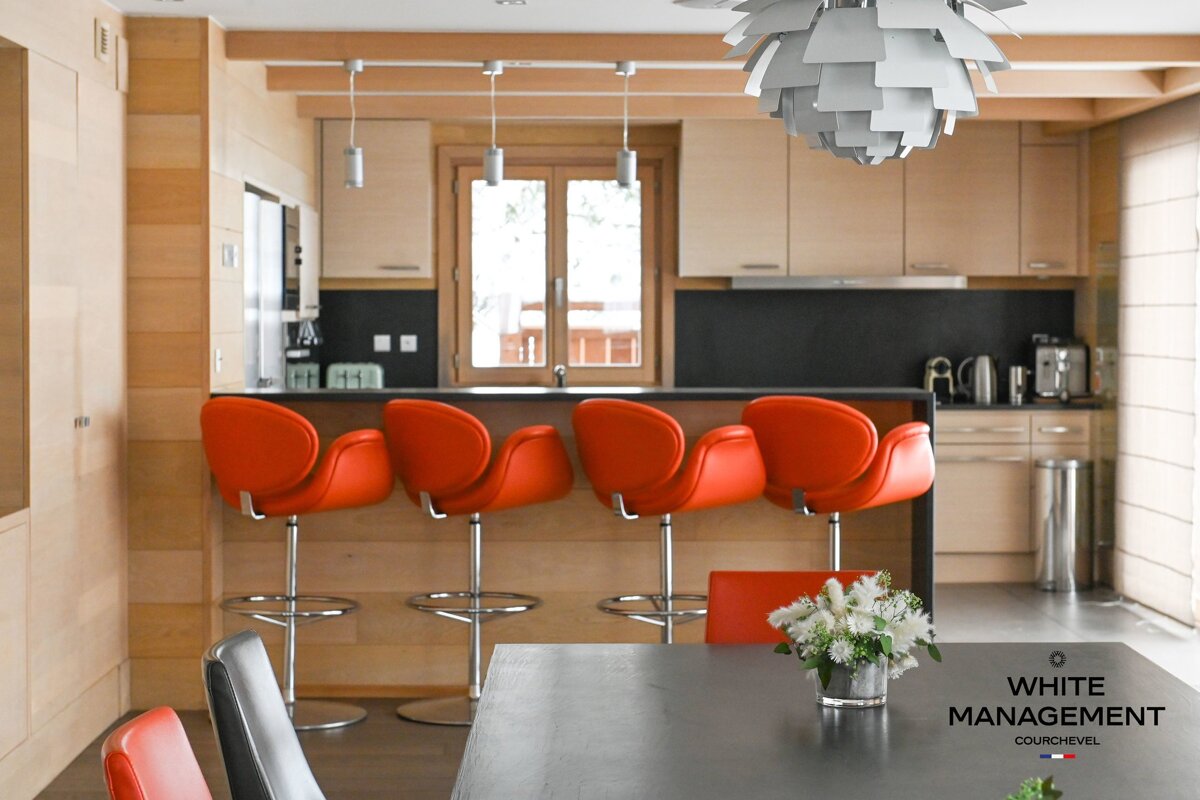 A modern wooden kitchen featuring four vibrant orange bar stools lined up at the counter, with a dining table in the foreground.