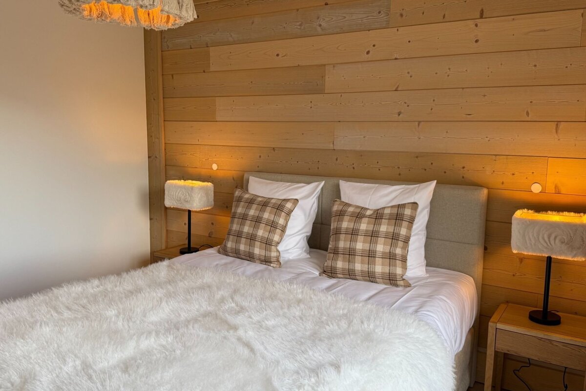 Cozy cabin bedroom featuring a wood-paneled wall, fluffy white bedding, plaid pillows, and warm glowing lamps.