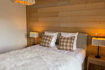 Cozy cabin bedroom featuring a wood-paneled wall, fluffy white bedding, plaid pillows, and warm glowing lamps.