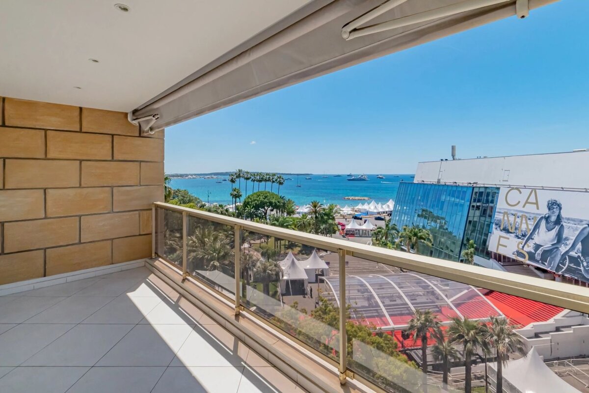 Balcony overlooking Cannes, with a blue sea, beach, palm trees, and the iconic Palais des Festivals under a clear sky.