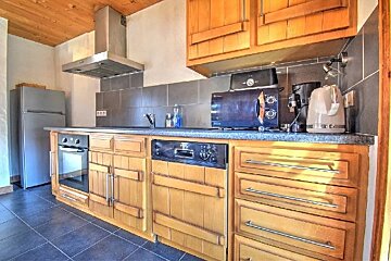 A rustic kitchen with wooden cabinets, a fridge, oven, microwave, coffee maker, and kettle. It features a dark tiled backsplash and countertop.