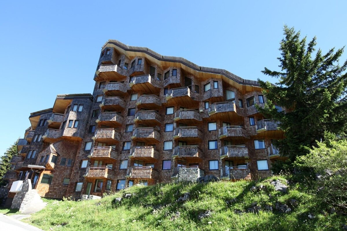 A modern apartment building with balconies stands amidst deep snow in a mountain village, backed by snow-covered peaks under a clear blue sky.